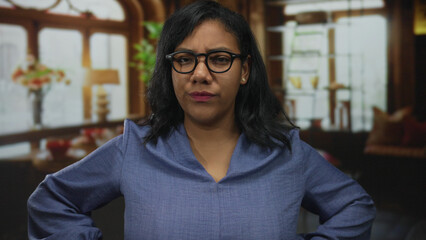 Woman in blue blouse with glasses and furrowed brow stands arms akimbo in building lit by daylight; annoyance.