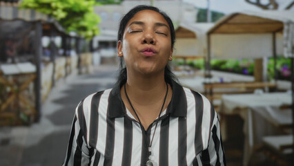 Referee woman claps hands on street terrace with whistle around neck and black and white striped shirt under leafy canopy; joy.