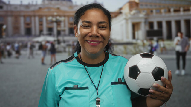 Woman referee holds soccer ball in front of building wearing turquoise referee jersey with visible whistle; confidence.
