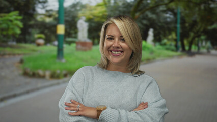 Obraz premium Woman smiles and crosses arms on paved street lined with green trees under soft daylight; confidence empowerment positivity selfesteem.