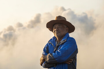 Portrait of elderly Asian man standing with arms crossed in industrial area with smoke rising from chimneys. Air pollution, environmental problems.