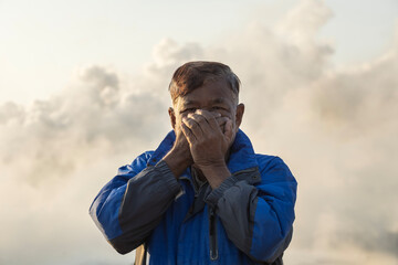 Air pollution: Portrait of elderly man covers his face and mouth, with smoke from factory chimney in the background.