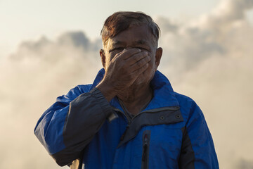 Asian elderly man covering his nose and mouth stands against backdrop of industrial smoke. air quality bad and pollution