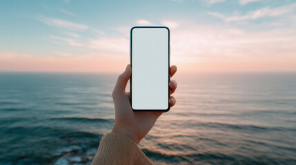 穏やかな海と空が広がる夕暮れ時（または朝焼け）の浜辺で、白い画面のスマートフォンを水平線に向けて掲げる手元、モックアップ