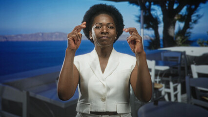 Fototapeta premium Woman pinching fingertips near face in white jacket, making a precise tiny gesture on a building terrace overlooking the sea; precision determination.