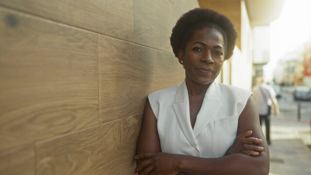Woman leaning against wooden wall with arms crossed on street, slight smile and relaxed posture; resilience confidence.