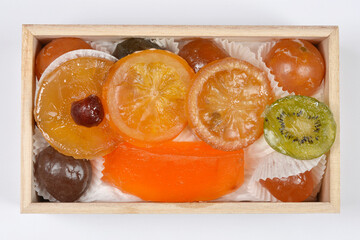 A top down view of various colorful candied fruits including orange slices kiwi and cherries neatly arranged in a small wooden box on a white background