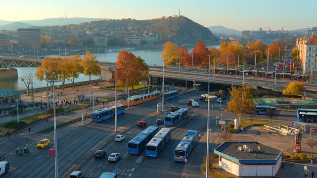 Panoramic aerial view of a busy Budapest street in autumn, featuring architecture and Petofi bridge, yellow fall trees, city traffic with cars and buses, and pedestrians walking on sunny sidewalks.
