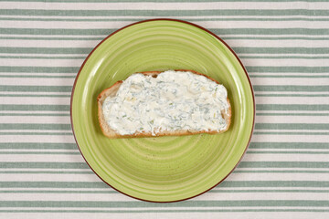 A top down view of a single slice of bread topped with a thick white spread of sour cream dill and garlic served on a green plate over a striped tablecloth