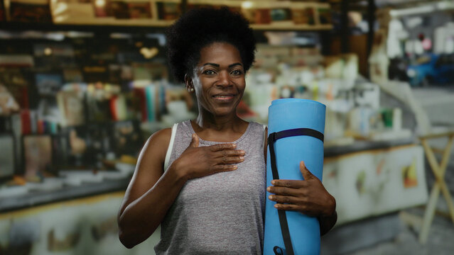 African american woman holding yoga mat outdoors near market, appearing calm and centered, wearing a casual tank top, suggesting a city street setting blending health and relaxation.