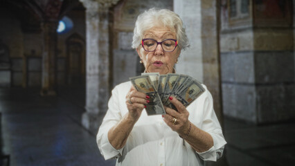 Senior woman counting banknotes and holding cash with visible hands in a stone building; surprise...