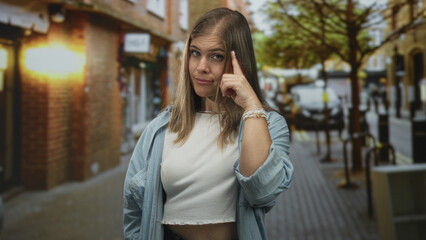 Blonde woman wearing denim shirt over white crop top pointing finger forward in busy city street; assertion confidence boldness attention.