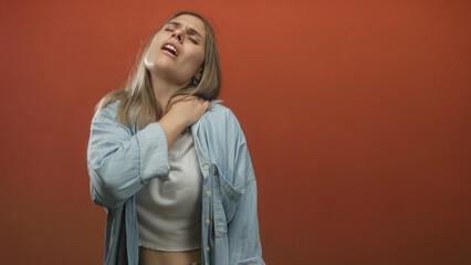 Young blonde woman wearing denim shirt over white top grips shoulder in studio with orange wall and...