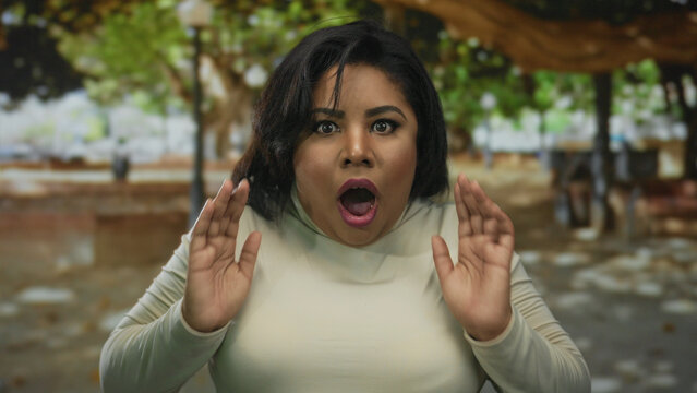 Young plus size woman with shocked expression in outdoor park setting under dappled tree shade displaying surprise and emotion