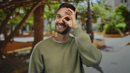 Fototapeta premium Hispanic man gesturing ok sign in a sunny park, surrounded by green outdoor scenery, expressing joyful playfulness with nature in the background.