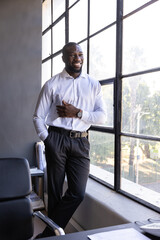 African American man leaning on windowsill wearing suit near office desk with documents, copy space