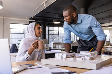 Diverse coworkers discussing architectural model beside laptop in open-plan office with coffee