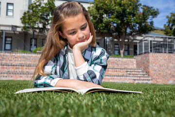 Child girl reading open book on green grass in courtyard in front of building