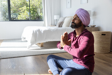 Mid adult indian man meditating pressing hands at chest in bedroom next to bed, window, copy space © wavebreak3