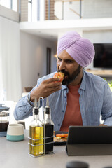 Mid adult indian man in lilac turban taking bite from tomato sandwich in kitchen, tablet © wavebreak3