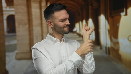Young man holding wrist with expression of pain standing in a historic university campus surrounded by arches suggesting an academic setting.