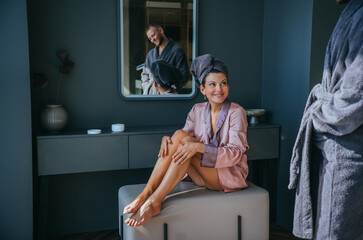 A woman sits on a vanity bench smiling at her partner, dressed in matching pink and grey bathrobes within a serene bathroom setting.