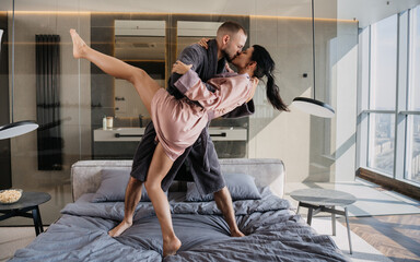 Playful couple in robes sharing a passionate dance on the bed, surrounded by modern design and warm lighting, capturing a candid moment of joy.