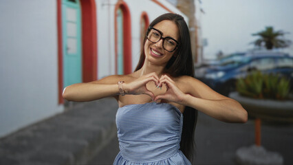 Woman forms heart with hands on street in front of colorful building wearing strapless blue dress and glasses, smiling at camera; love friendship.