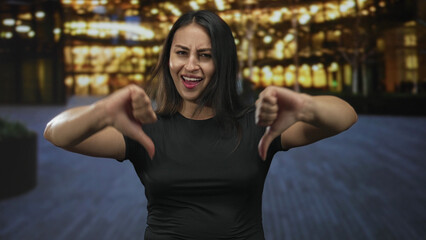 Young hispanic woman giving thumbs down with both hands and a grimaced face in front of a lit...