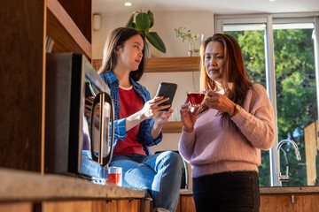 Asian mother and daughter chatting by kitchen island holding smartphone and cup of tea