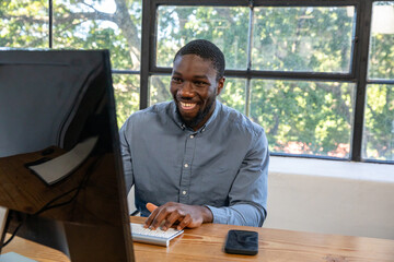 African american man typing on keyboard at office desk by window with monitor, smartphone