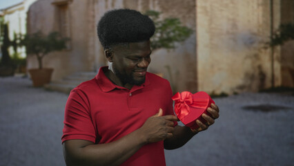 Man in red polo holds red heart gift box and points finger to box with both hands on old town stone street near building entrance; romance surprise.