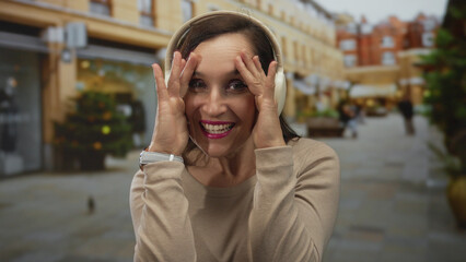 Fototapeta premium Young hispanic woman with white headphones covers face with both hands on busy downtown street; playfulness joy curiosity.