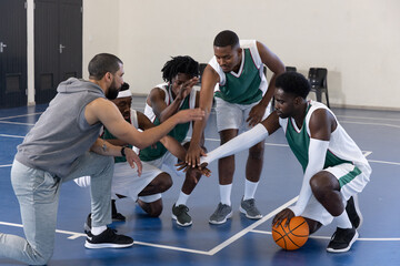 Male coach and male basketball players placing hands on basketball on blue court in jerseys