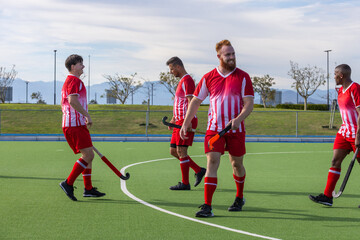 Diverse male teammates walking across synthetic turf field hockey pitch with field hockey sticks
