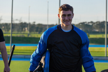 Fototapeta premium Teenage boy standing on synthetic hockey pitch in blue-black goalkeeper gear holding helmet