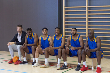 Diverse male basketball team sitting on bench in sports hall holding basketball, water bottle