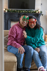 Naklejka premium Smiling diverse senior couple in holiday tree and antler glasses sitting on couch by fireplace