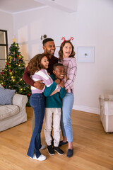 Obraz premium Diverse family hugging in living room with mother wearing holiday headband, christmas tree