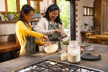 Diverse female friends wearing aprons whisking batter and adding cherries in home kitchen with bowl