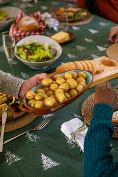 Diverse friends in sweaters passing brown ceramic dish of potatoes at home dining table