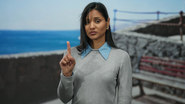 Woman gesturing on seaside promenade with ocean background, indicating no with finger, young latin, wearing grey sweater, beach setting creating a relaxed outdoor atmosphere.