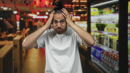 Young arab hispanic man holding head with both hands in busy restaurant building; stress shopping mishap.