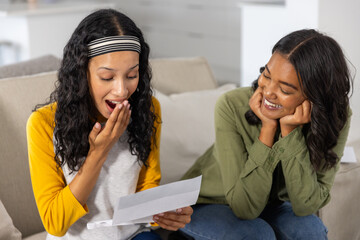Diverse mom and teenage daughter sitting on beige sofa in living room reading letter together