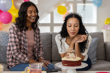 Diverse mom and teenage daughter celebrating 18th birthday on grey sofa in living room with cake