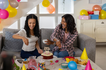 Diverse mom and teenage daughter celebrating birthday on couch in living room with cake, balloons