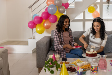 Diverse mom and teenage daughter slicing red velvet birthday cake amid balloons, party snacks