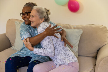 Senior female friends hugging on beige couch in living room with pastel balloons and confetti