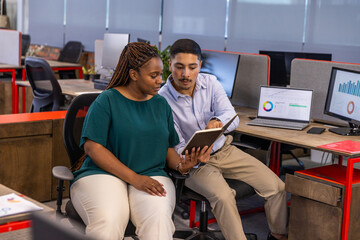 Diverse coworkers reviewing notebook in open-plan office with laptop graphs, smartphone