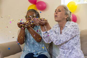 Diverse senior female friends blowing paper confetti while sitting on couch at home with balloons
